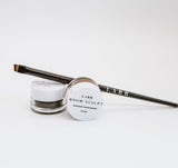 Black precision angle brush balanced on a jar of brow sculpt waterproof shaping pomade. A second brow sculpt eyebrow product is balanced on its side in front of the brush.