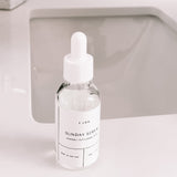 A clear and white bottle of Sunday Serum sitting on the edge of a white counter next to a sink. 
