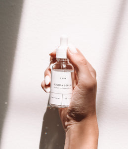 A hand holding a clear and white bottle of Sunday Serum with shadowed and refracted light in the image