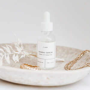 A clear and white bottle of Sunday Serum in a stone tray next to a gold ring and necklace with a dried plant stem. 