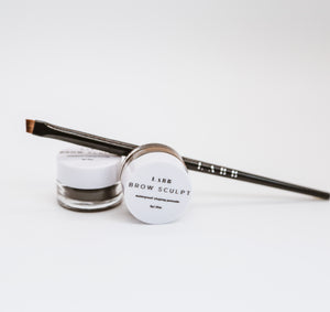 Black precision angle brush balanced on a jar of brow sculpt waterproof shaping pomade. A second brow sculpt eyebrow product is balanced on its side in front of the brush. 